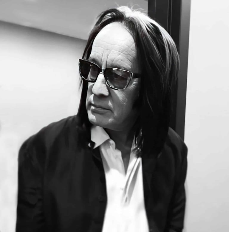 Man with long dark hair and sunglasses wearing a black jacket and white shirt in black and white portrait.