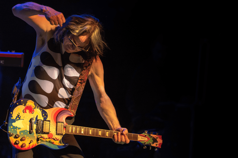 Musician with long hair playing yellow electric guitar under blue and orange stage lighting.