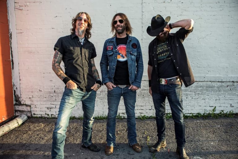 Three musicians in rock attire stand against a weathered white brick wall in an industrial setting.