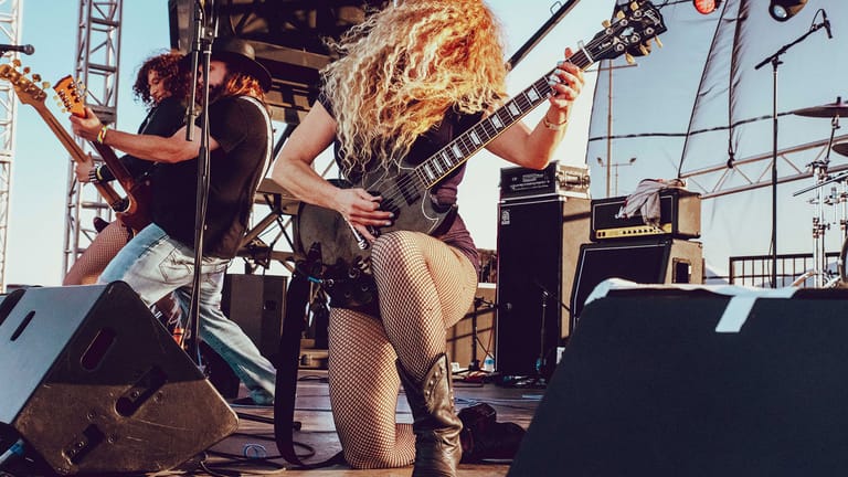 Guitarist with long blonde hair performing energetically on stage under bright sunlight with band members and equipment visible behind.