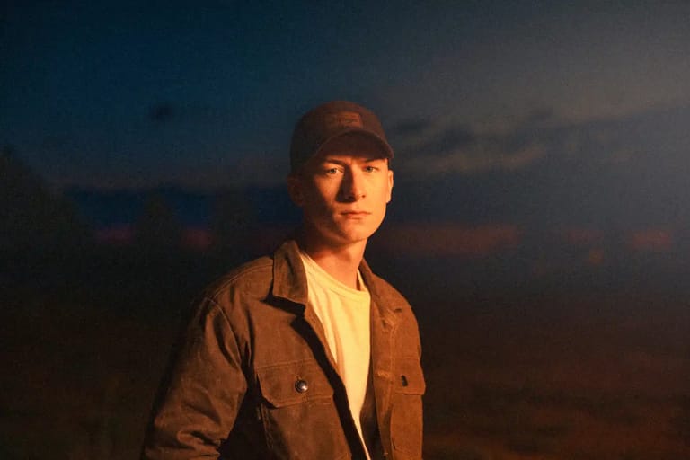 Person in brown jacket and cap standing outdoors at golden hour against a twilight sky.