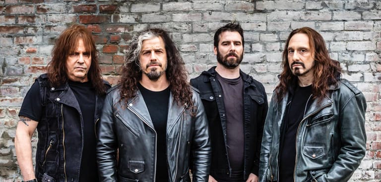Four long-haired male musicians in black leather jackets stand against a weathered brick wall.