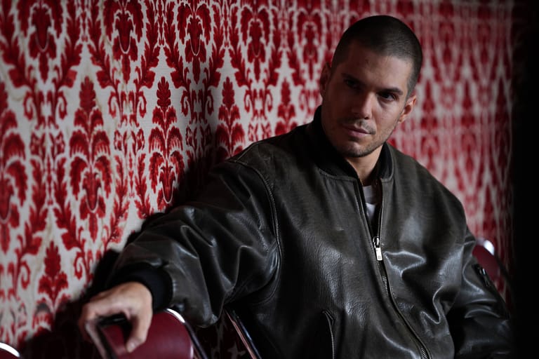 Man in black leather jacket seated against red and white patterned wallpaper backdrop.