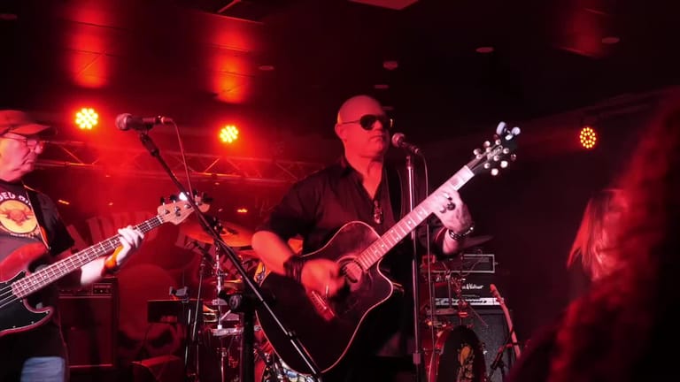 Guitarist performs under red stage lighting whilst drummer plays in the background.