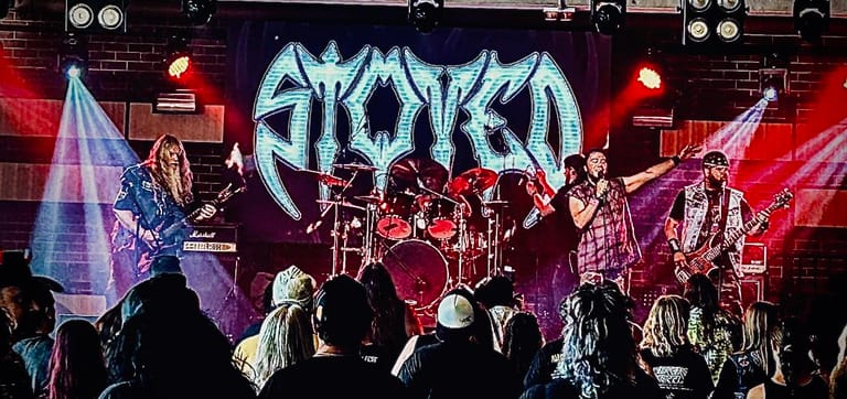 Five-piece band performing on stage beneath blue "AMORZA" lettering with red and purple stage lighting.
