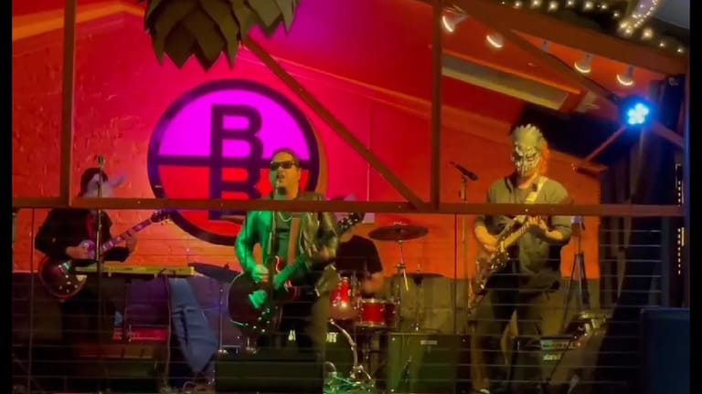Band performs on stage illuminated by pink and orange lighting, with a large glowing "B" logo backdrop.