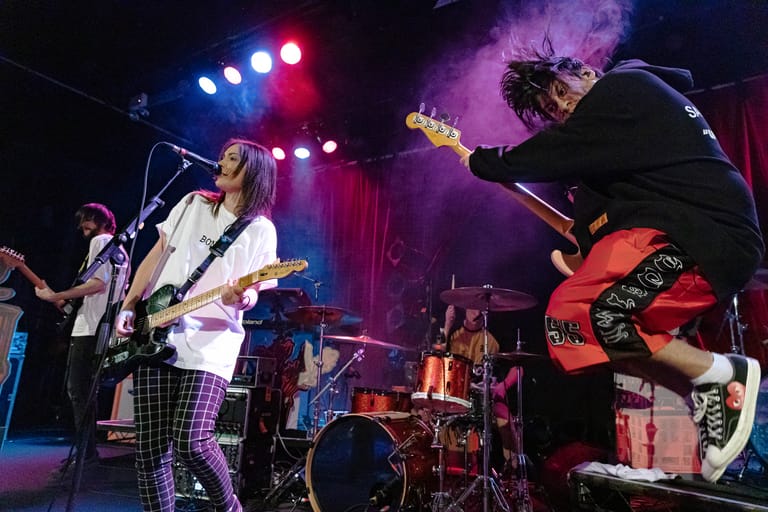 Band performs live on stage with guitarist in white jacket and drummer silhouetted against purple stage lighting.