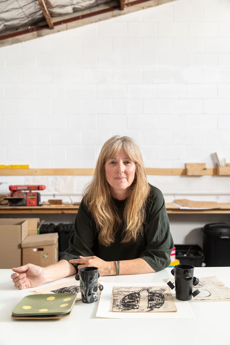 Woman with long blonde hair seated at a workshop table surrounded by design materials and tools.