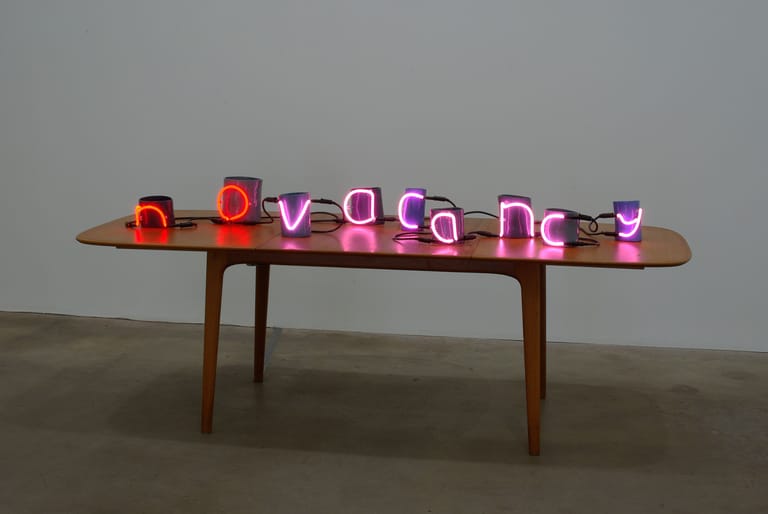 Purple neon lettering spelling "vacancies" displayed on a wooden mid-century table.