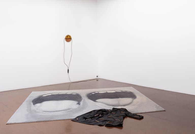 Wooden sphere suspended above white paper with two circular cutouts and coiled black rope on gallery floor.