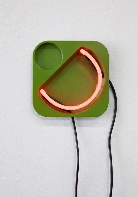 Neon smiley face glowing in pink on a green square panel against white wall.
