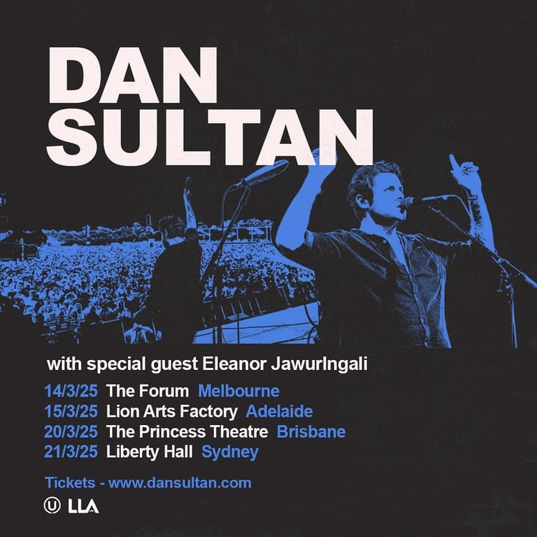 Dan Sultan performing on stage with raised fist, illuminated in blue stage lighting against a crowd backdrop.