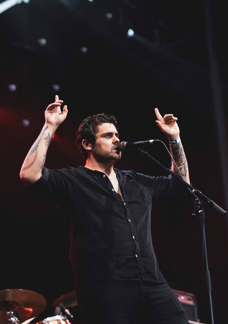 Male performer in black shirt at microphone with both arms raised, stage lights and bokeh background.
