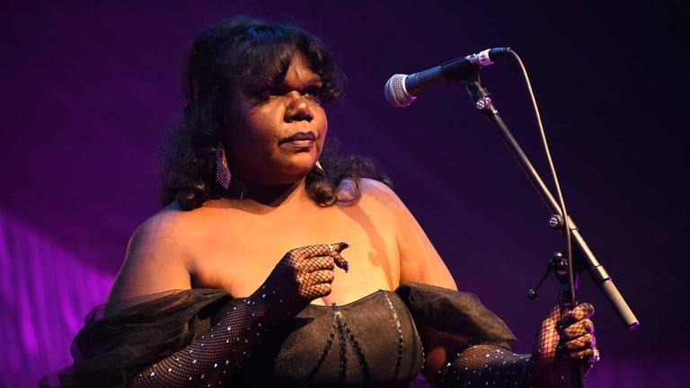 Singer in black gloves and off-shoulder top performing at microphone under purple stage lighting.