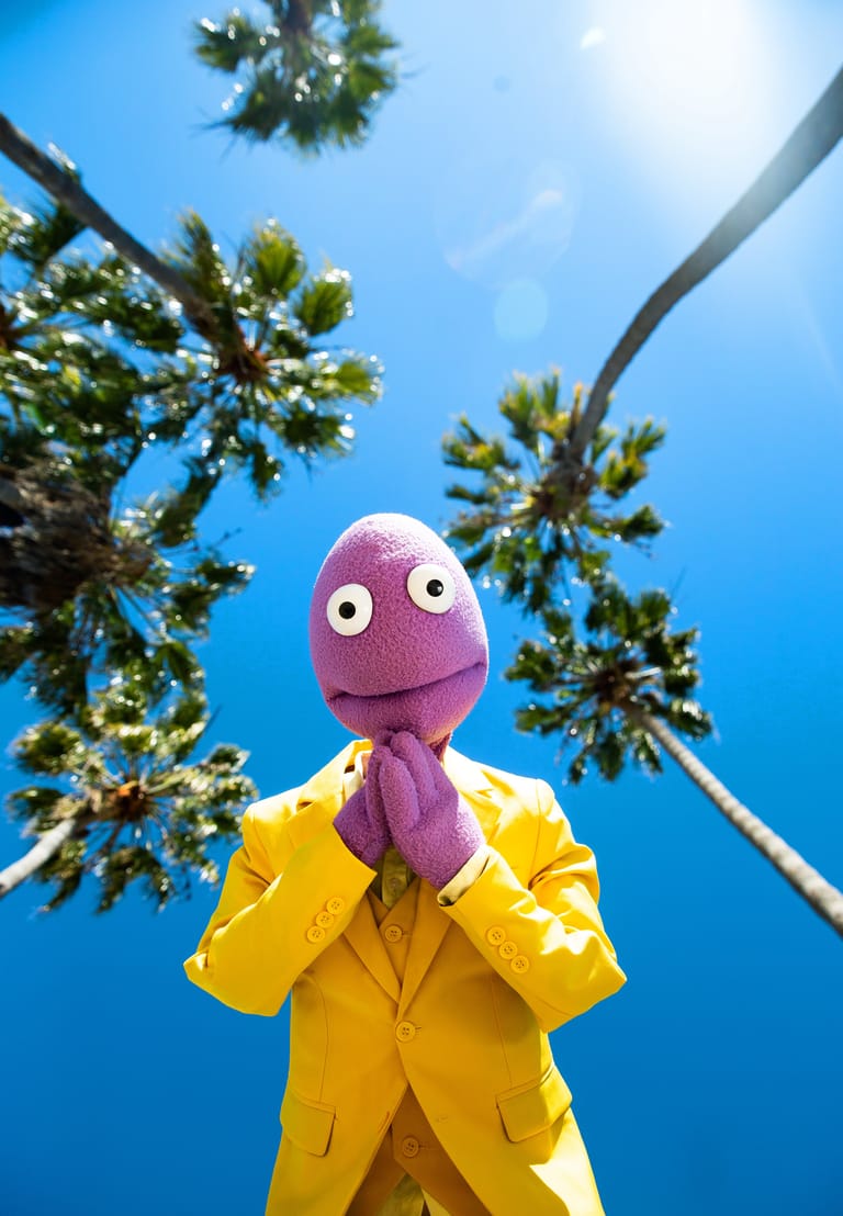 Purple puppet character in bright yellow jacket against tall palm trees and blue sky.