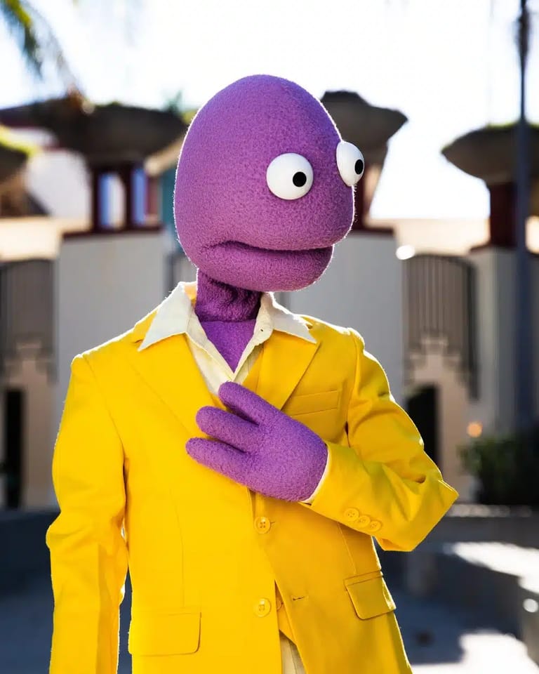 Character in vibrant purple head and yellow jacket stands outdoors against modern architecture backdrop.