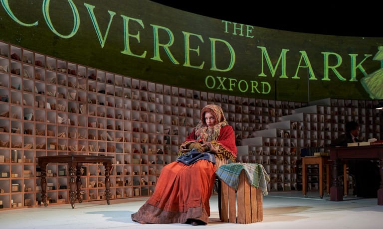 Actor in red dress seated on stage against brick wall backdrop during theatre production.