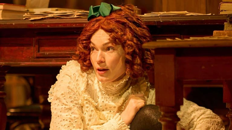 Performer with curled auburn hair and cream lace costume against ornate red interior set.