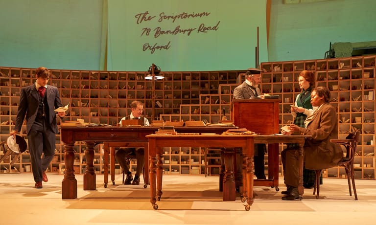 Four performers positioned around a wooden table on a stage set with terracotta tiled walls during a theatrical production.