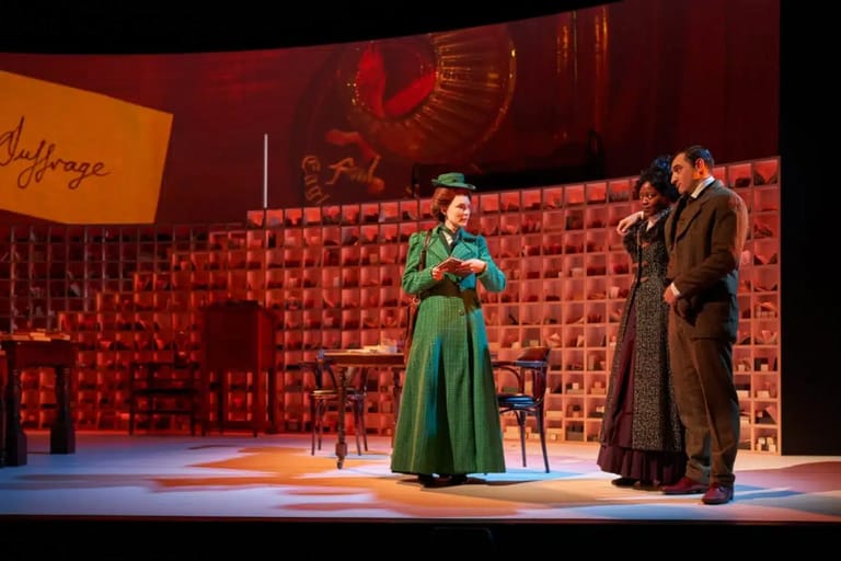 Woman in green dress and two figures in dark coats on stage set with red brick backdrop and warm lighting.