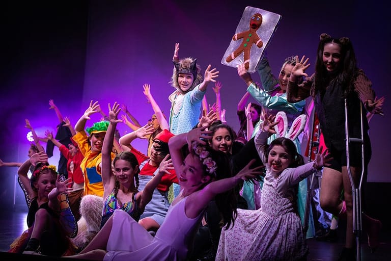 Ensemble of performers in colourful costumes on stage bathed in purple lighting, arms raised expressively.