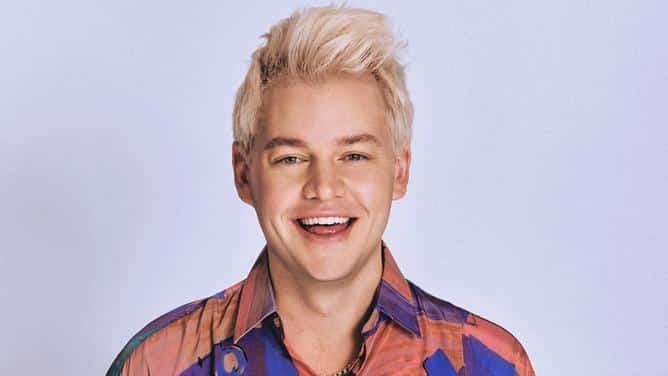 Person with blonde hair wearing a blue and orange patterned shirt against a lavender background.