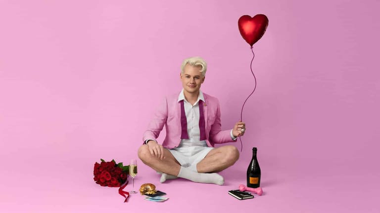 Person in pink blazer holding red heart balloon, seated cross-legged against pink background with flowers and wine bottle.