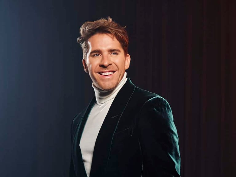 Man in black velvet jacket and white shirt smiling against dark blue backdrop.
