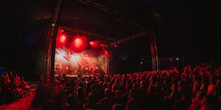 Large outdoor stage bathed in red light with crowd of spectators facing the illuminated performance platform at night.