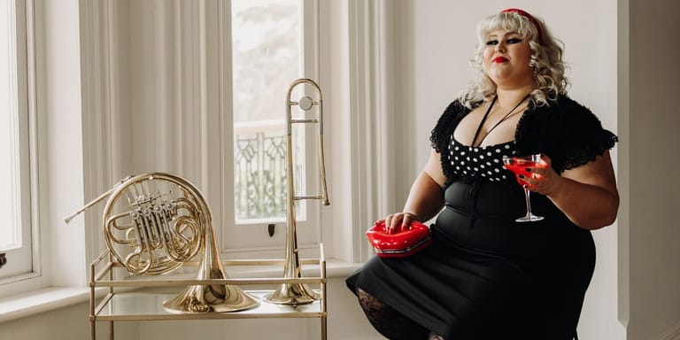 Woman in black dress holding a cocktail sits beside brass musical instruments by a sunlit window.