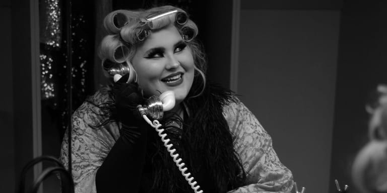 Woman in vintage-style curled hair and patterned jacket speaks into a telephone in black-and-white photograph.