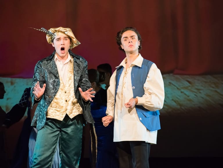 Two performers in period costume strike exaggerated poses on stage against a deep red theatre curtain.