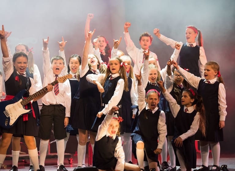Group of young performers in black and white school uniforms with fists raised on stage, celebrating together under warm stage lighting.