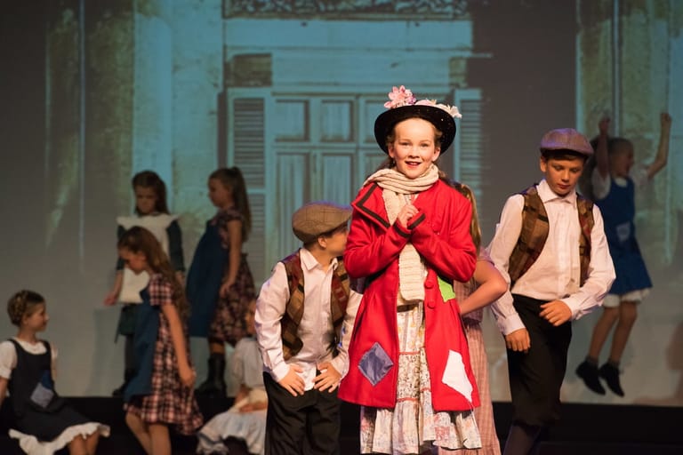 Child in red coat and hat performs centre stage with costumed ensemble cast in period dress behind.