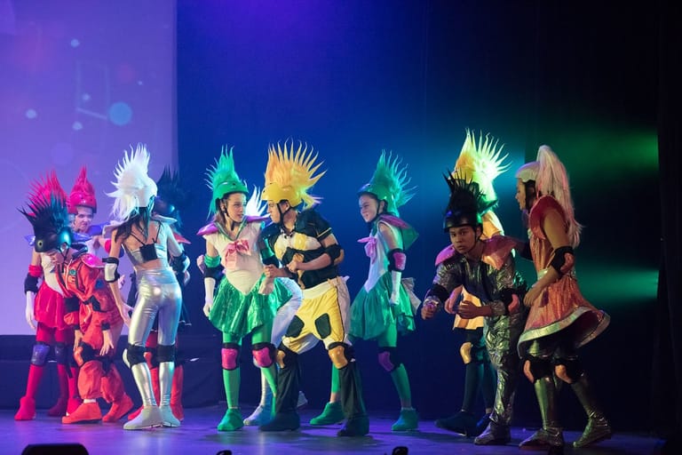 Performers in vibrant anime-style costumes with spiky hair stand posed together on a stage lit by purple and green lights.