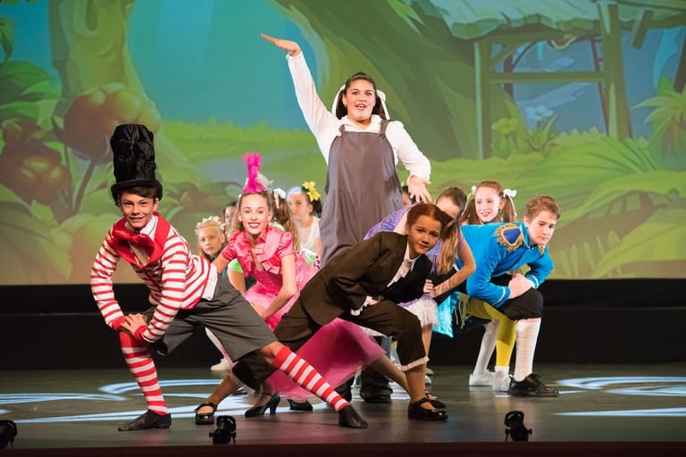 Group of young performers in colourful costumes dancing on stage with jungle scenery backdrop.