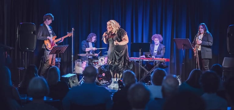 Band performs on stage with blonde female vocalist centre, surrounded by musicians and blue curtain backdrop, audience silhouettes in foreground.