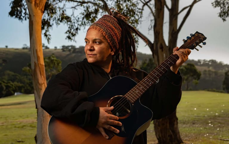 Musician holding an acoustic guitar in a rural paddock setting with eucalyptus trees.