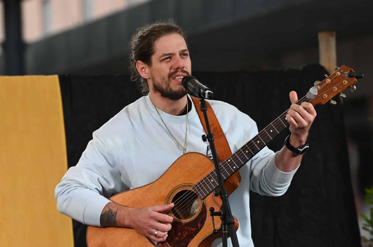 Musician performing with acoustic guitar and microphone on stage, wearing a light blue shirt.