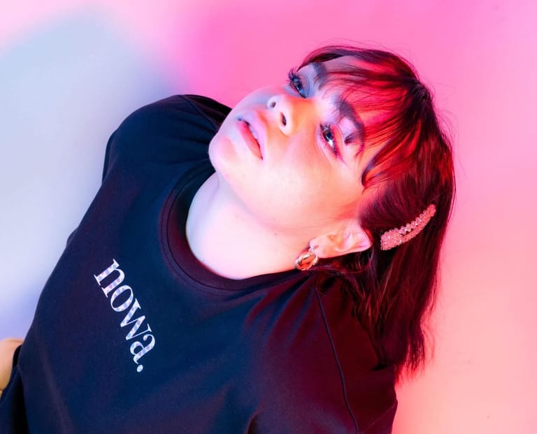 Person with red hair and purple eyeshadow tilting head back against pink and blue gradient background.