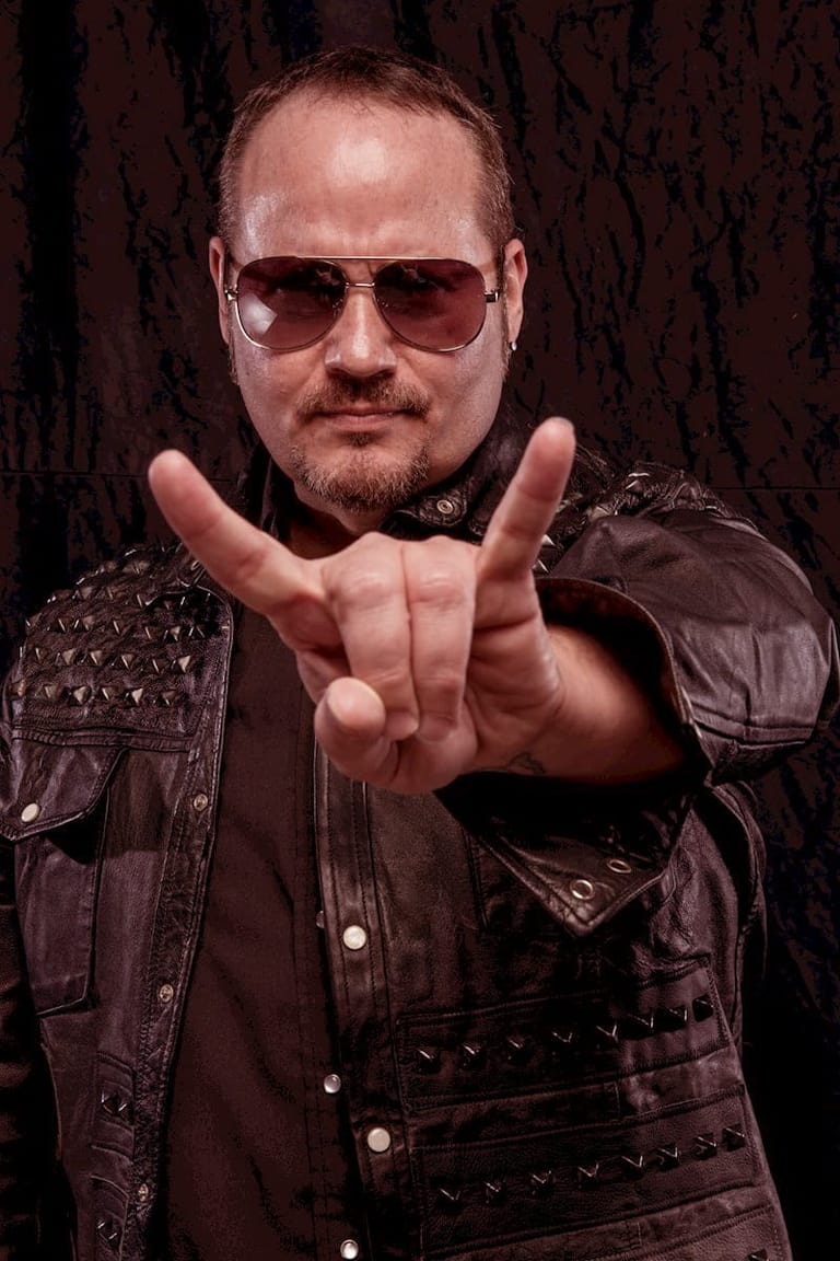 Man in studded leather jacket and aviator sunglasses making a rock gesture against dark textured backdrop.