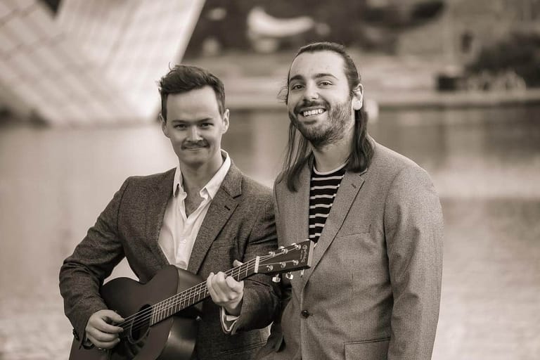 Two musicians smile while holding an acoustic guitar in a sepia-toned portrait.