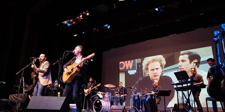 Band performing on stage with projected image of historical figure on large screen backdrop.