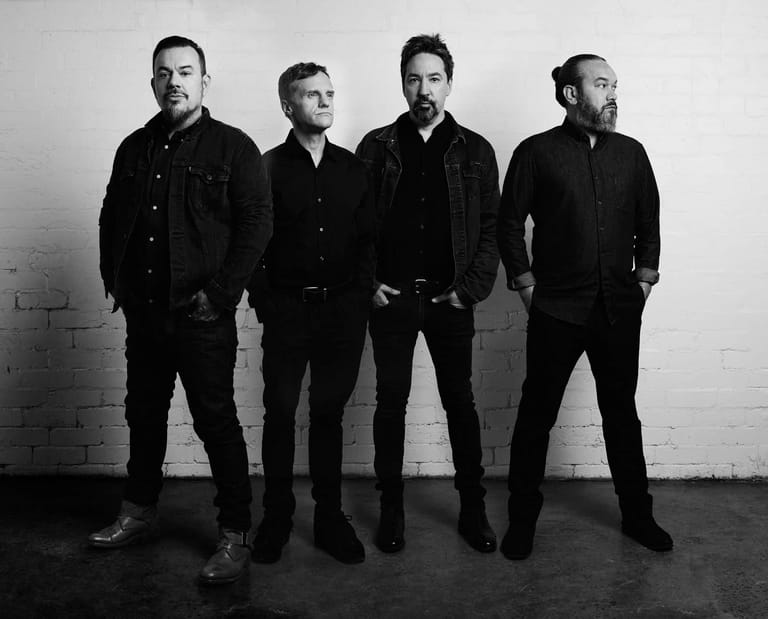 Four men in dark suits stand against a white brick wall in a black and white promotional portrait.