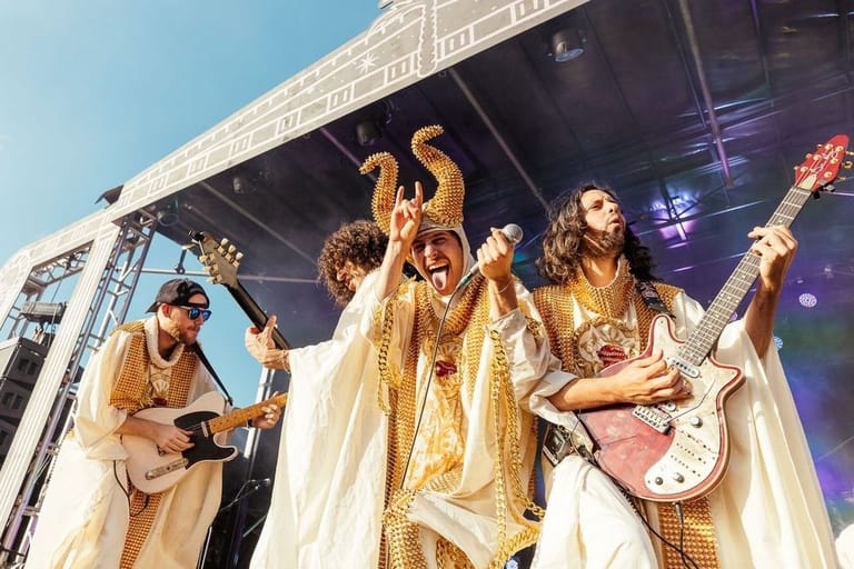 Band members in gold and cream robes perform with guitars on an outdoor festival stage under clear blue sky.