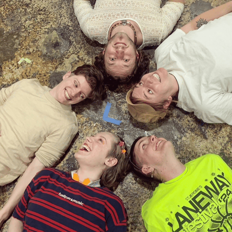 Five young people lying on concrete ground with heads together, smiling at camera.