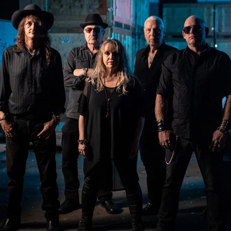 Five band members in dark clothing stand posed together against a blue-lit industrial backdrop.