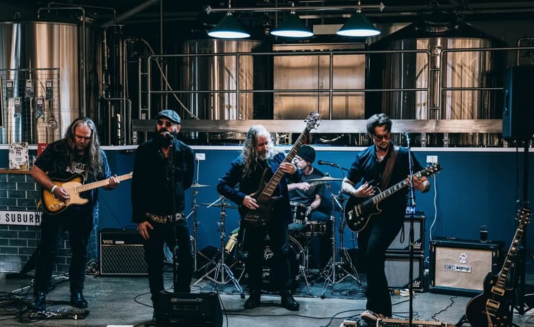 Five musicians perform on stage in a brewery setting, with blue walls and industrial brewing equipment visible behind them.