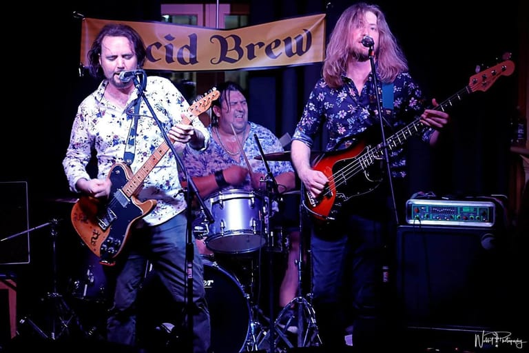 Two guitarists and a drummer perform on stage beneath an "Acid Brew" banner in a dimly lit venue.