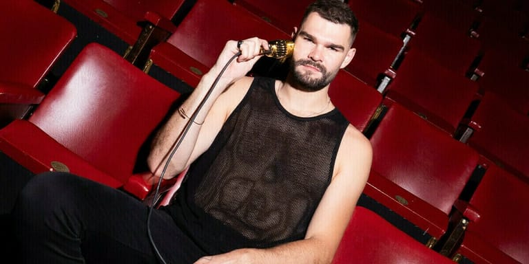 Man in sleeveless black shirt reclines in red theatre seat, holding a jacket over his shoulder.